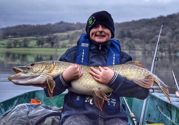 a fisherman on his boat holding a giant northern pike that he has caught on a cloudy day with moderate temperatures