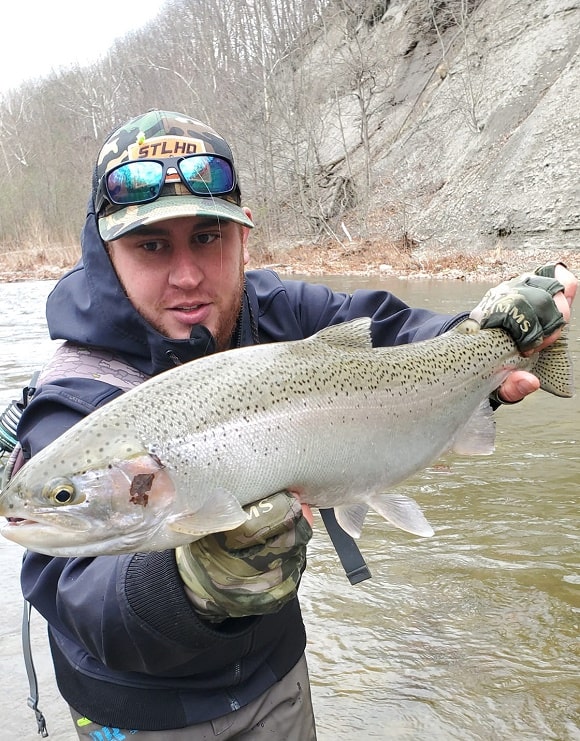a trout angler on a river holding a big steelhead that he has caught on a streamer