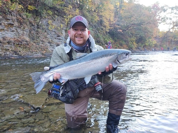 a happy angler standing in a shallow river and holding a really big steelhead trout