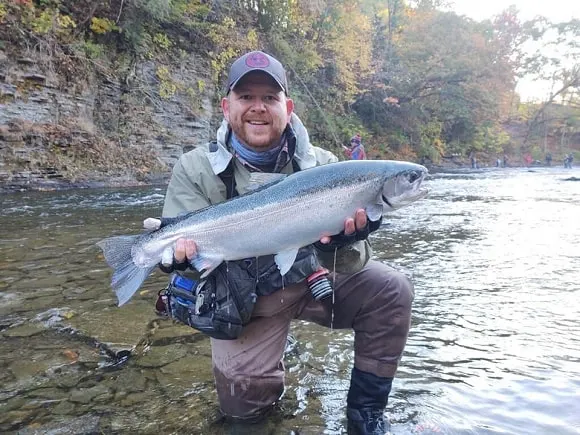 a happy angler standing in a shallow river and holding a really big steelhead trout