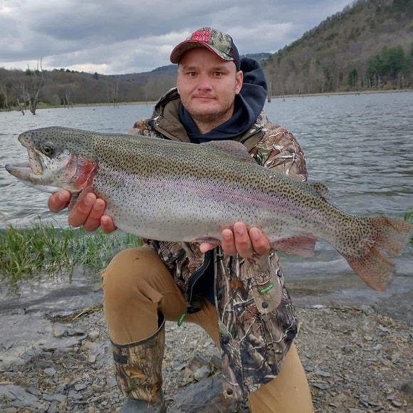 a trout angler in front of a lake holding a big rainbow trout