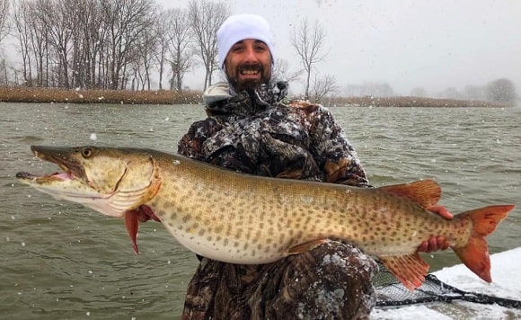 a US angler on his boat holding a big musky that he has cuahgt in a snow storm