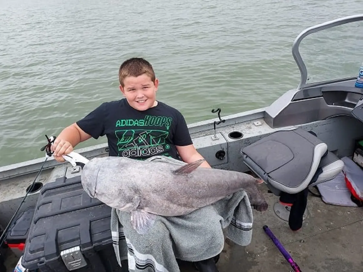 a young catfish angler on a boat holding a giant blue catfish